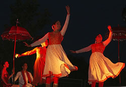 Three femal Kathak dancers with skirts flying and raised left arms. Bride and groom seated in the background on the left.